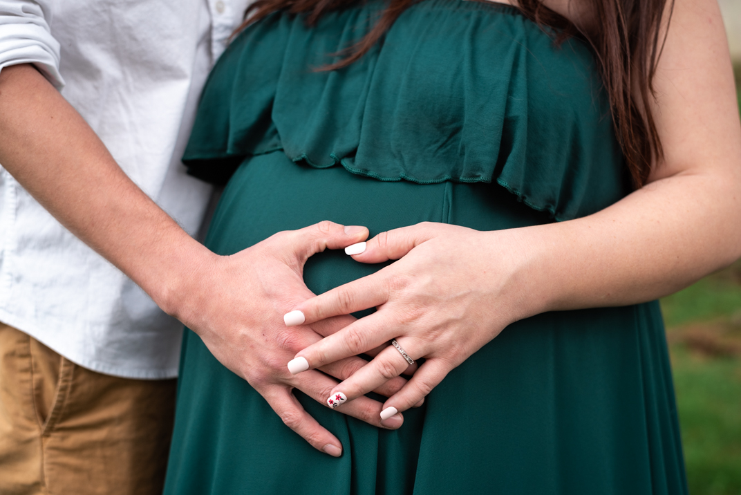 Maternity photo of a woman in a green dress with her hand on her stomach. Her husbands hand rests beside hers. This photo is the cover photo for a blog post and gallery of images related to this couple's maternity session in Bristol, Tennessee.