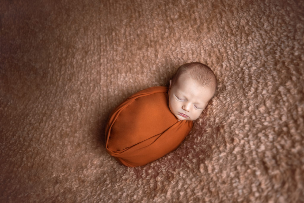 A caucasian newborn baby laying on a brown, fuzzy blanket and wrapped tightly in an orange swaddle cloth. The baby is asleep. The photograph was taken at a photography studio near Johnson City, Tennesseee.