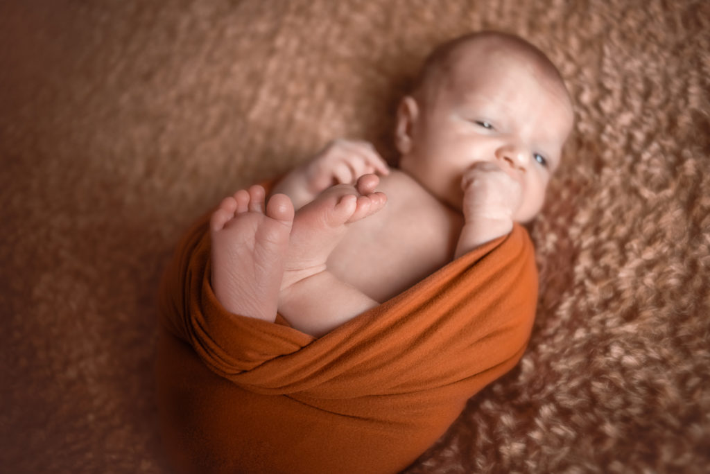 A caucasian newborn baby laying on a blanket and wrapped in an orange cloth. The photo is focused on the baby's feet and shows the details of the toes. The photograph was taken at a photography studio near Johnson City, Tennesseee.