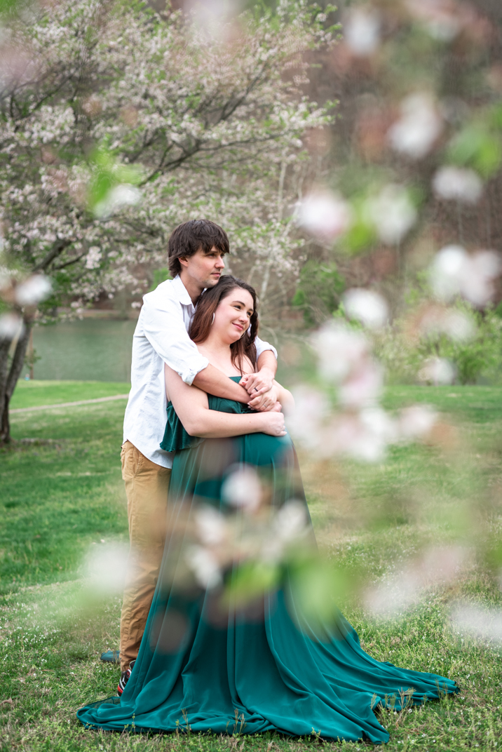 A man wraps his arms around a pregnant woman wearing a green gown. It's spring and they are surrounded by cherry blossoms.